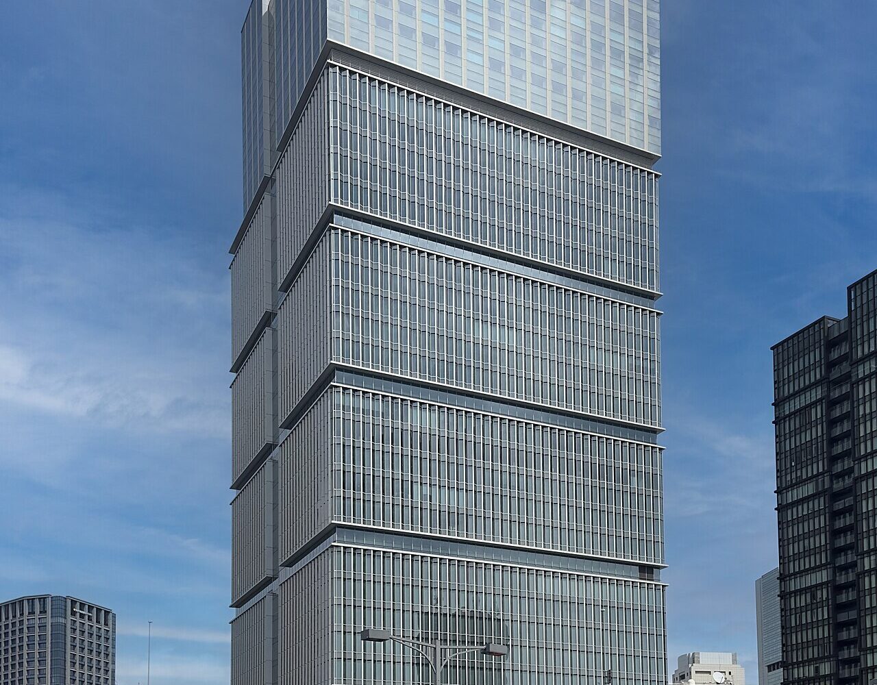 A tall glass and stone office tower rising above a tree-lined plaza in central Tokyo.