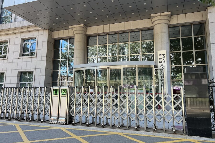 A retractable metal gate drawn across the covered entrance of a stone-clad government office building, with a vertical Chinese nameplate beside the revolving door.