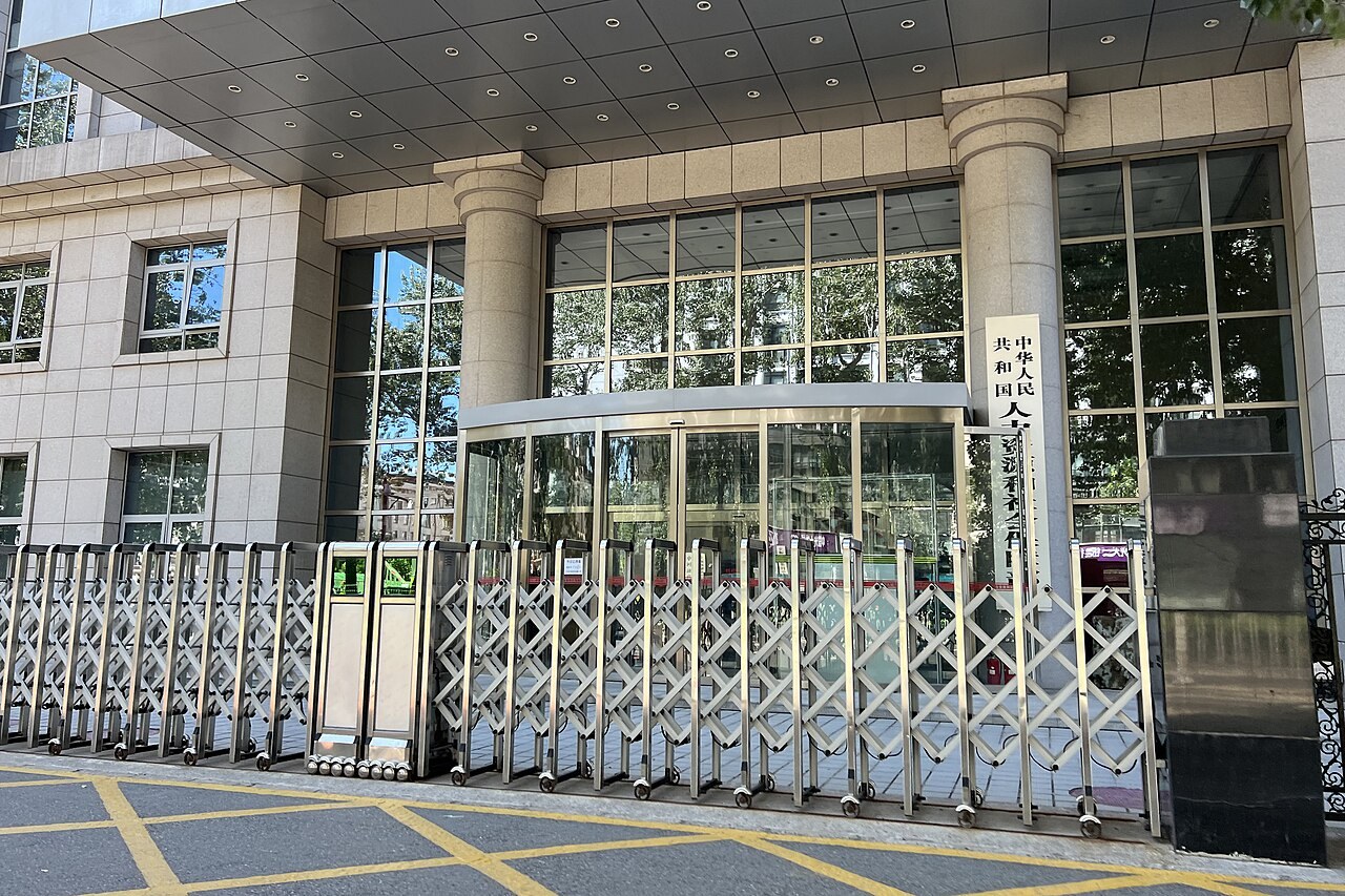 A retractable metal gate drawn across the covered entrance of a stone-clad government office building, with a vertical Chinese nameplate beside the revolving door.