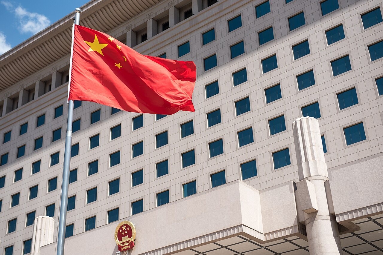China's national flag flying in front of the pale stone facade of the August 1st Building in Beijing, with the national emblem above the entrance