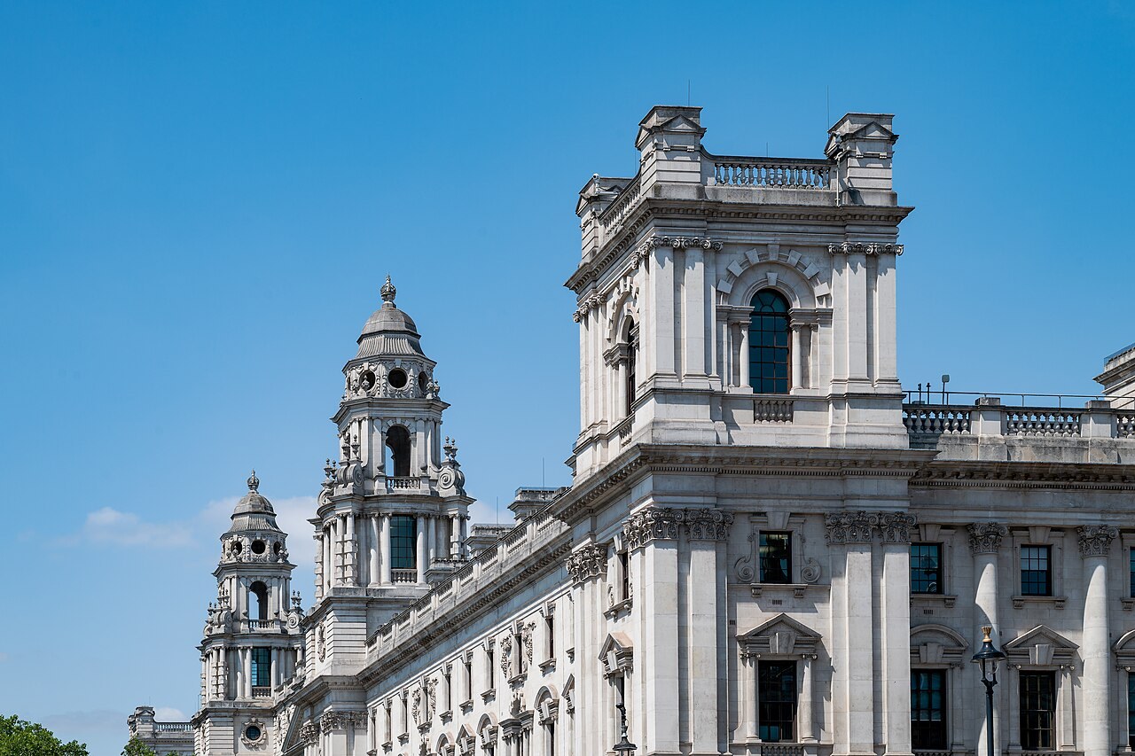 The upper storeys and corner towers of a large white Portland stone government building in Whitehall under a clear blue sky.