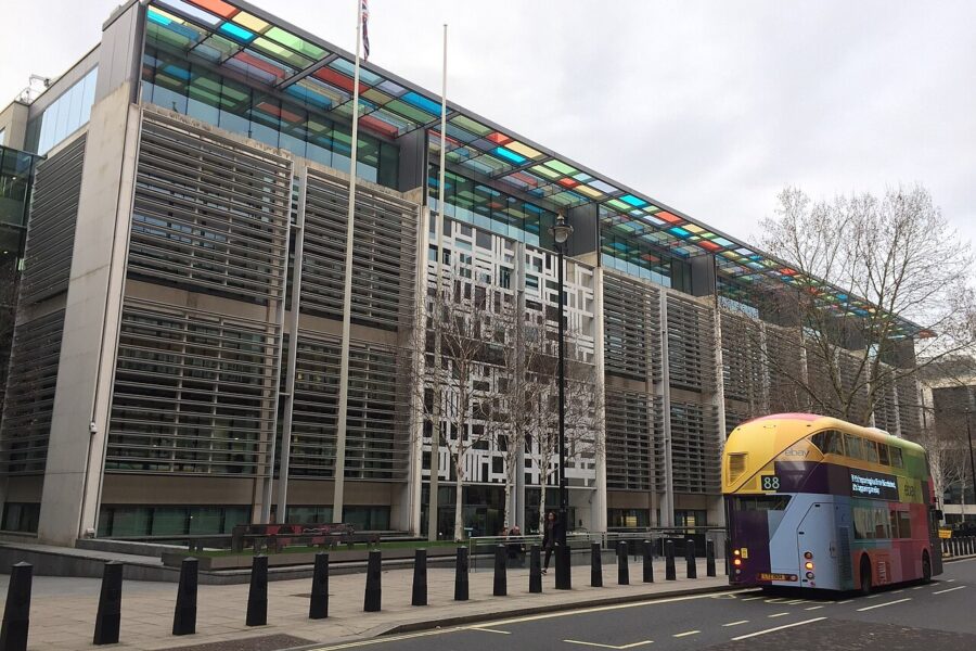 The long glass and louvred facade of the Home Office building on Marsham Street in London, with coloured glass panels along its roofline and a red double-decker bus passing on the road