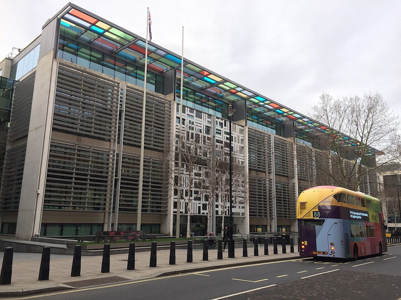 The long glass and louvred facade of the Home Office building on Marsham Street in London, with coloured glass panels along its roofline and a red double-decker bus passing on the road