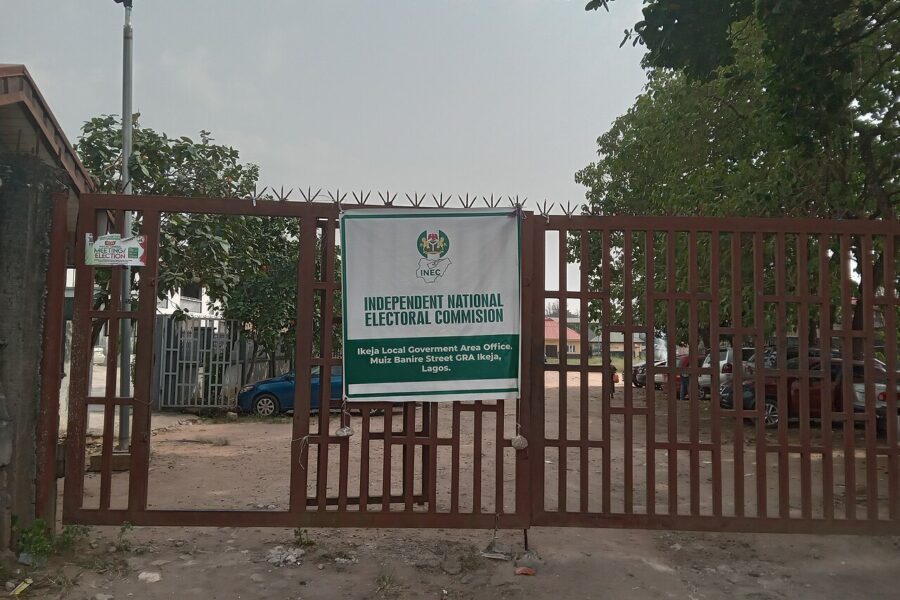 A closed red metal gate with a green and white Independent National Electoral Commission sign for the Ikeja local government area office, trees and parked cars behind it.