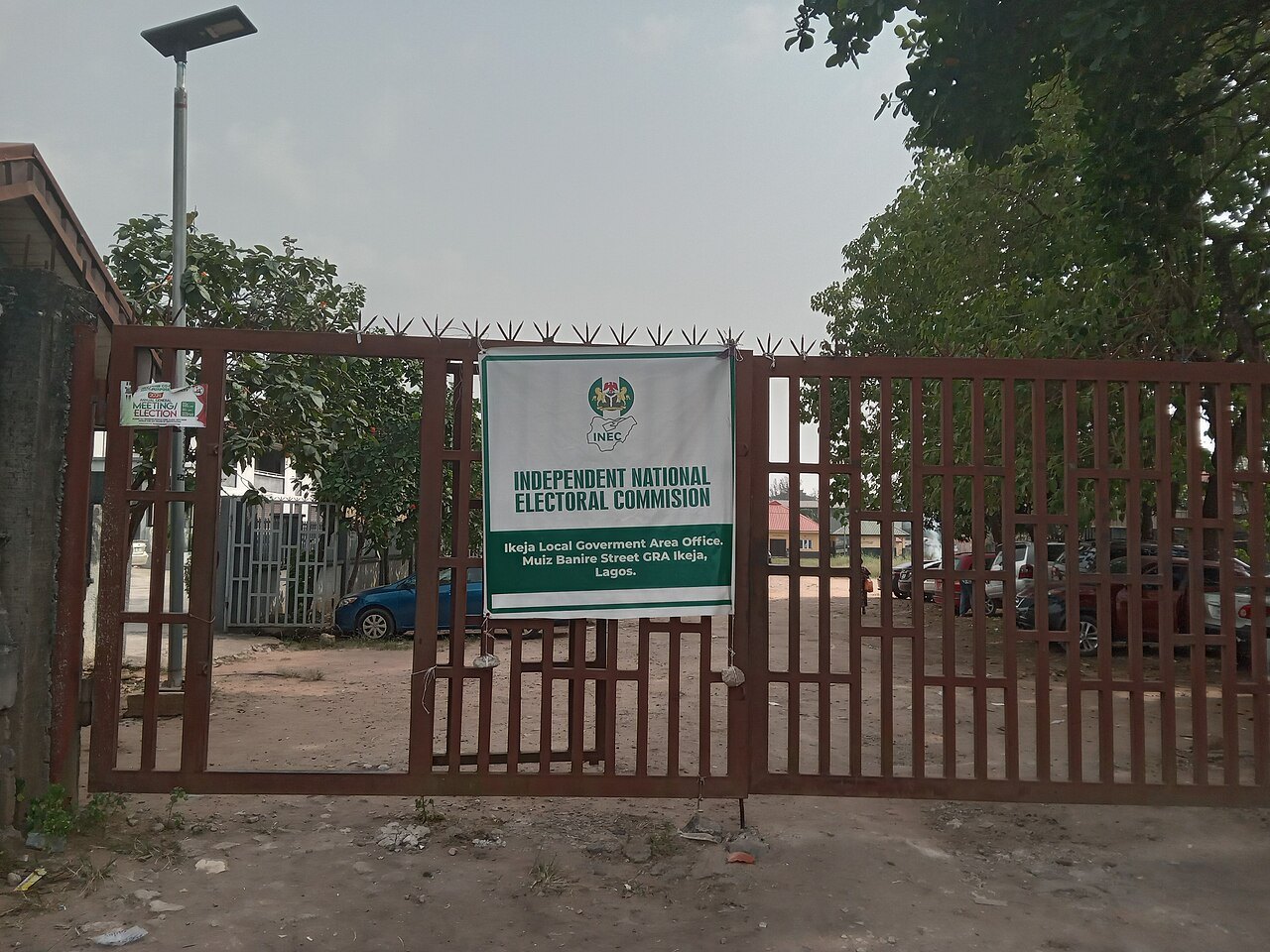 A closed red metal gate with a green and white Independent National Electoral Commission sign for the Ikeja local government area office, trees and parked cars behind it.