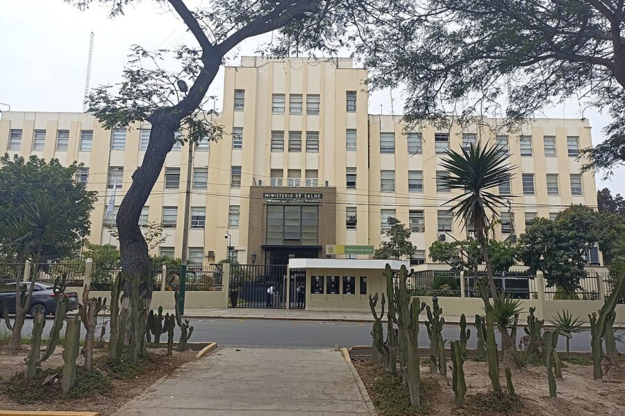 A cream-coloured multi-storey government building with a sign reading Ministerio de Salud above its gated entrance, framed by trees and cacti.