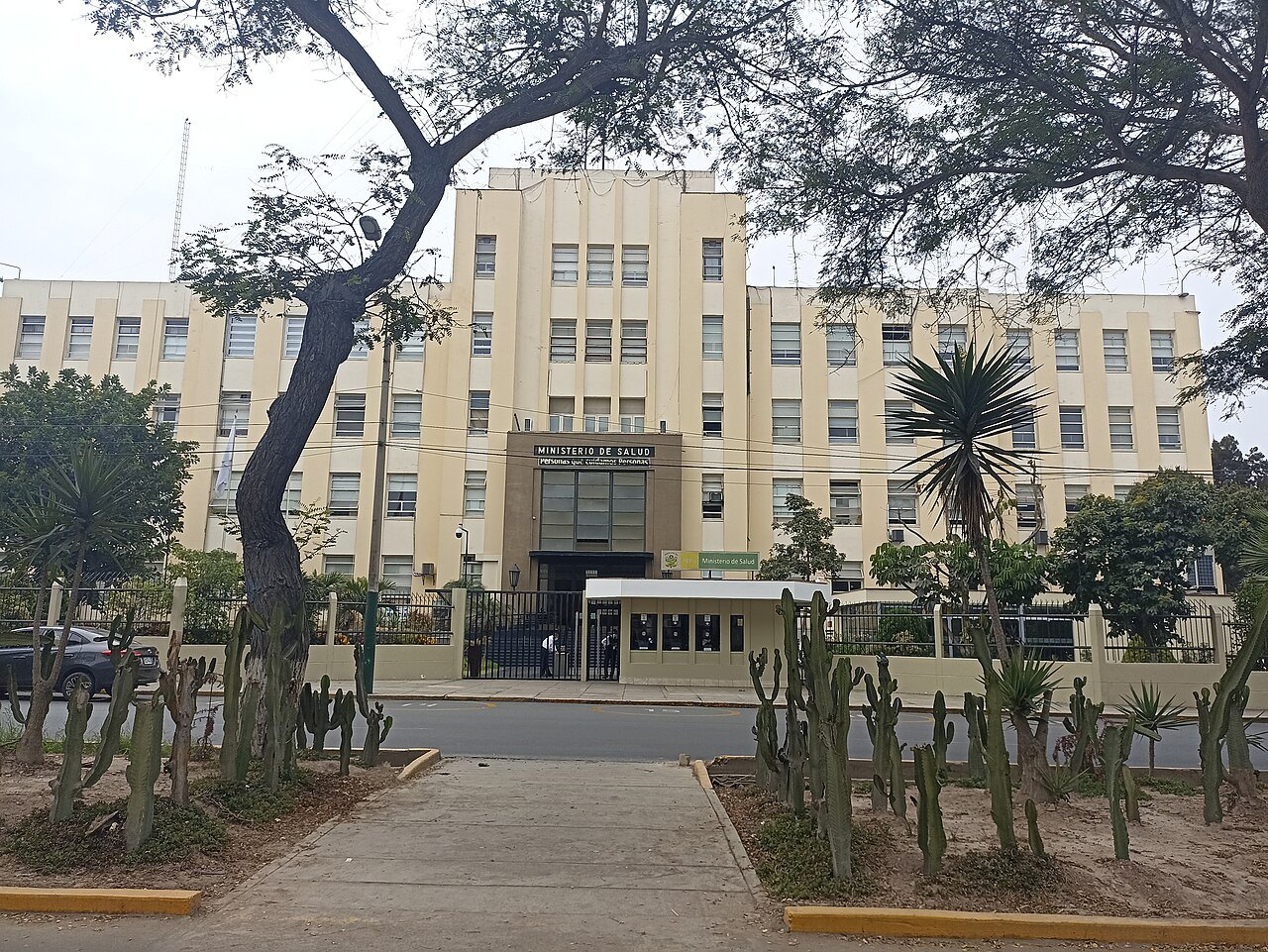 A cream-coloured multi-storey government building with a sign reading Ministerio de Salud above its gated entrance, framed by trees and cacti.