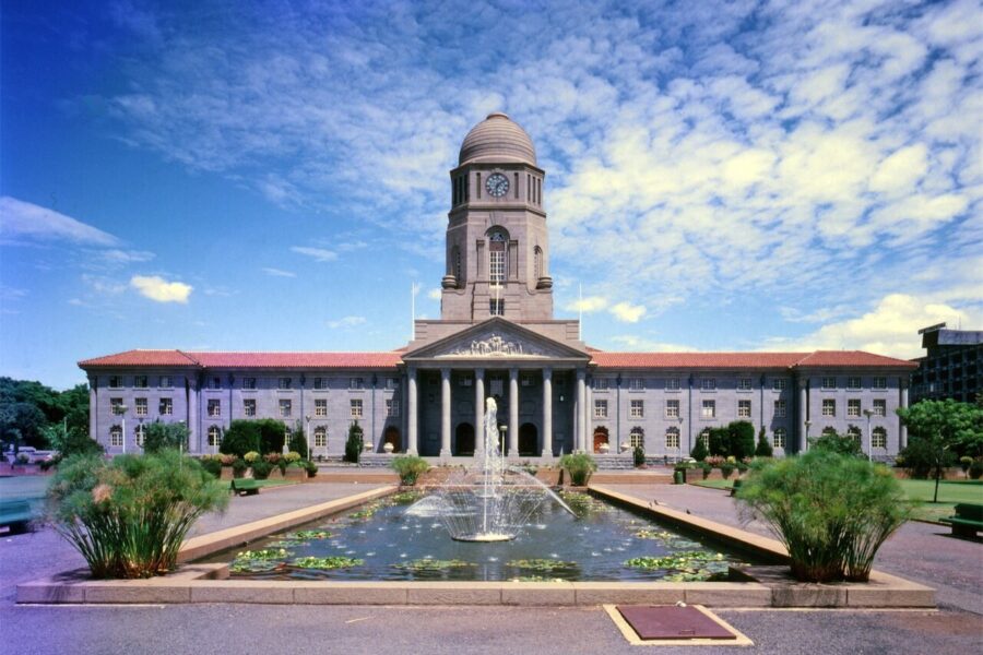 A symmetrical stone city hall with a central clock tower and a columned portico, seen across a long rectangular pond with a fountain.