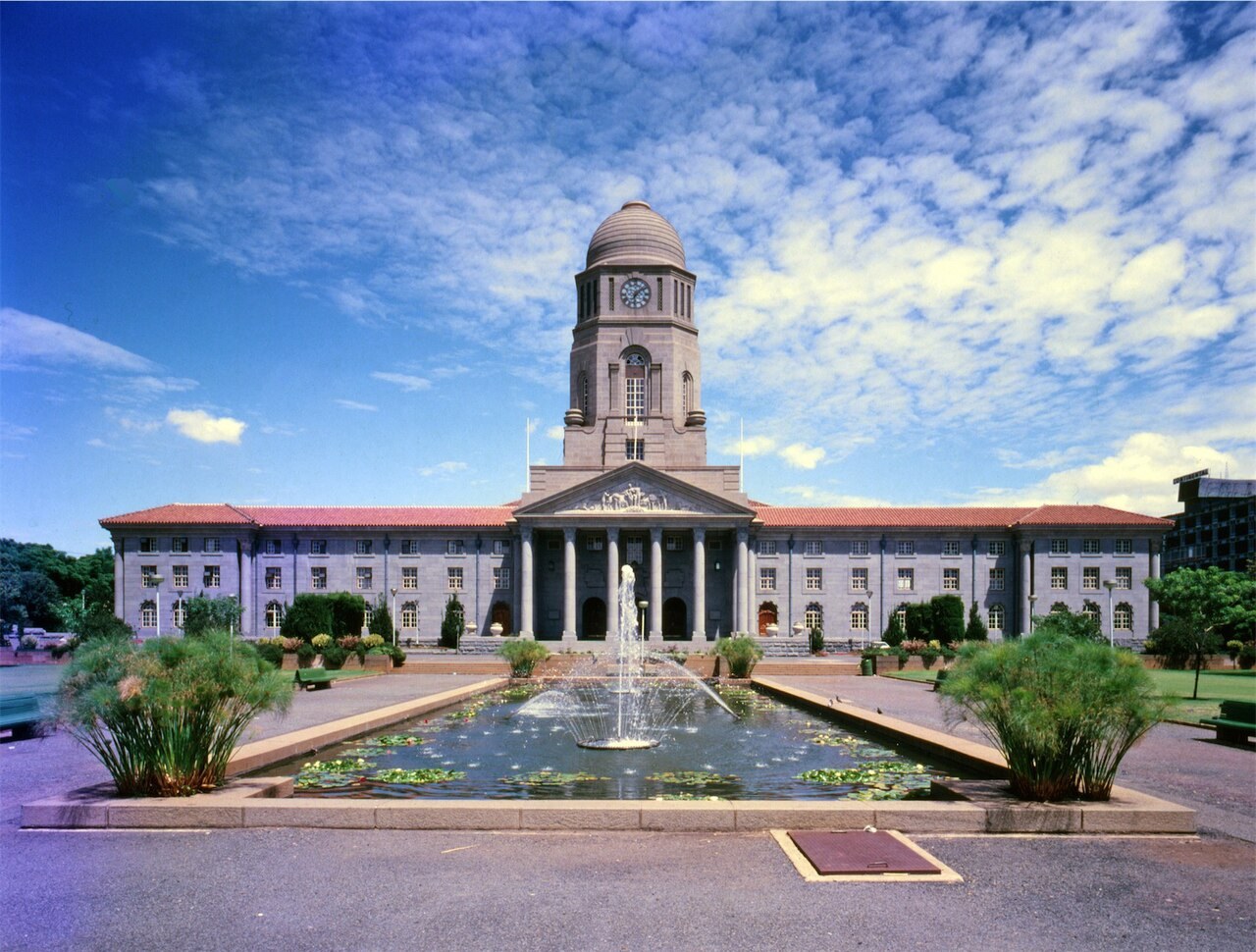 A symmetrical stone city hall with a central clock tower and a columned portico, seen across a long rectangular pond with a fountain.