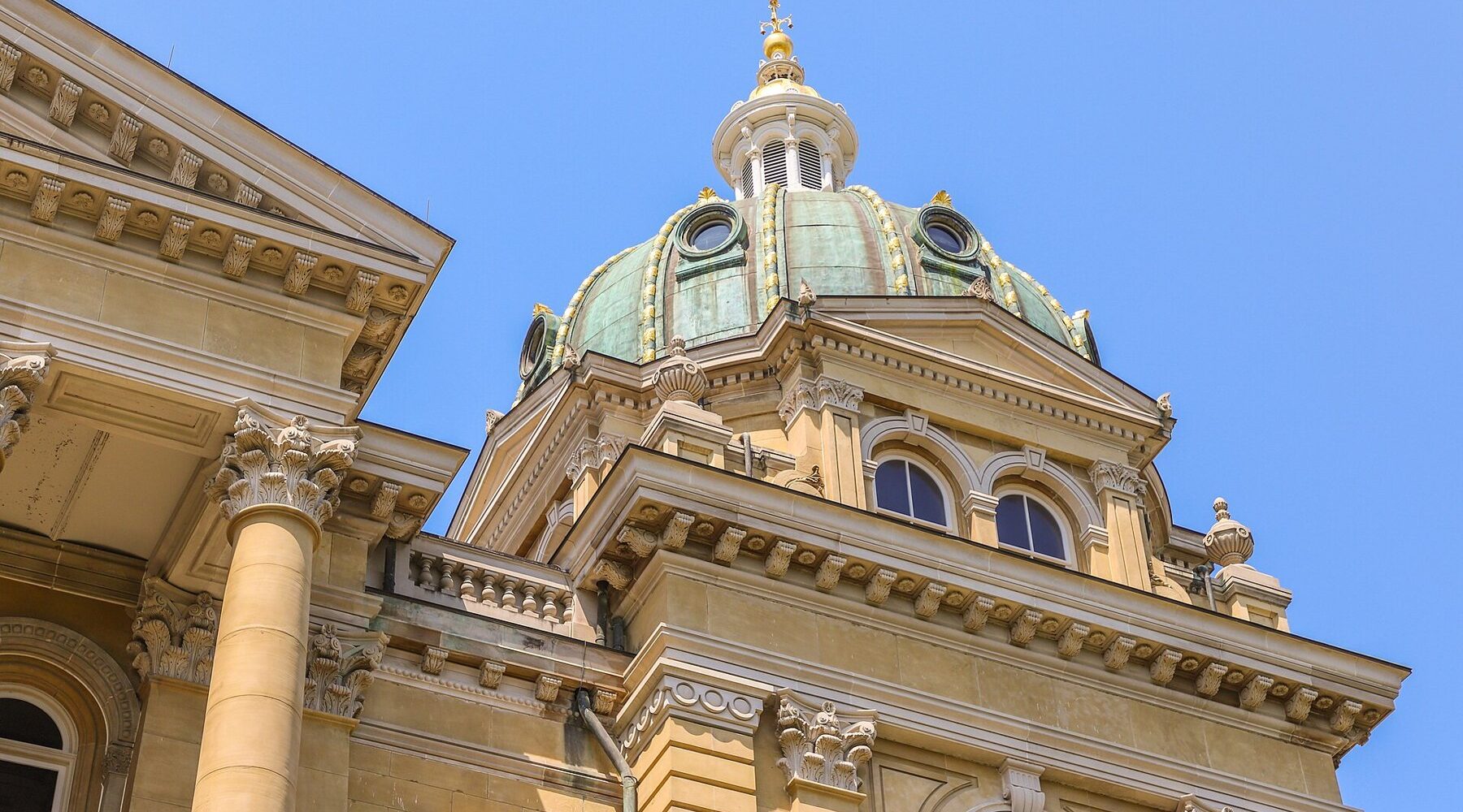 A green copper dome with gilded ribs on the Iowa State Capitol, rising above carved stone columns against a clear blue sky