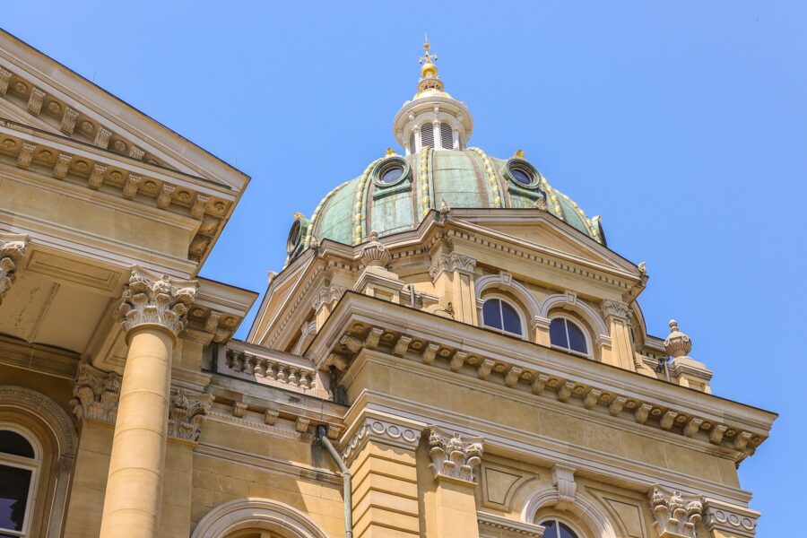 A green copper dome with gilded ribs on the Iowa State Capitol, rising above carved stone columns against a clear blue sky